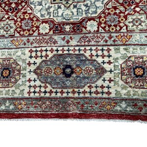 Puede incluir: Primer plano de una secci&oacute;n de una alfombra tradicional con un patr&oacute;n detallado. La alfombra presenta un fondo color crema con dise&ntilde;os intrincados en rojo, azul y dorado. El borde es de color rojo intenso y el dise&ntilde;o general es sim&eacute;trico.