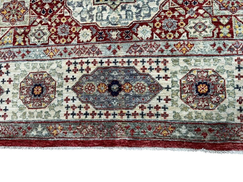 Puede incluir: Primer plano de una secci&oacute;n de una alfombra tradicional con un patr&oacute;n detallado. La alfombra presenta un fondo color crema con dise&ntilde;os intrincados en rojo, azul y dorado. El borde es de color rojo intenso y el dise&ntilde;o general es sim&eacute;trico.