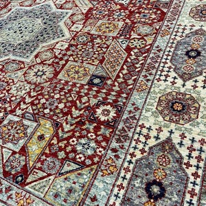 Puede incluir: Primer plano detallado de una gran alfombra ornamentada con un complejo patr&oacute;n geom&eacute;trico. La alfombra presenta una rica paleta de colores rojo, beige, gris y amarillo. El dise&ntilde;o incluye formas florales y geom&eacute;tricas repetidas.