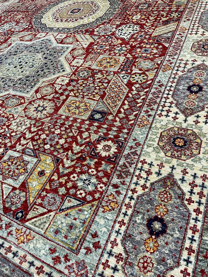 Puede incluir: Primer plano detallado de una gran alfombra ornamentada con un complejo patr&oacute;n geom&eacute;trico. La alfombra presenta una rica paleta de colores rojo, beige, gris y amarillo. El dise&ntilde;o incluye formas florales y geom&eacute;tricas repetidas.