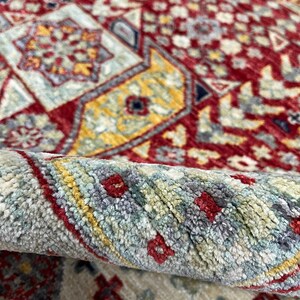 Puede incluir: Primer plano de una alfombra enrollada con un patr&oacute;n intrincado. La alfombra presenta un fondo rojo intenso con dise&ntilde;os geom&eacute;tricos en amarillo, azul y crema. El borde enrollado revela la textura y las variaciones de color.