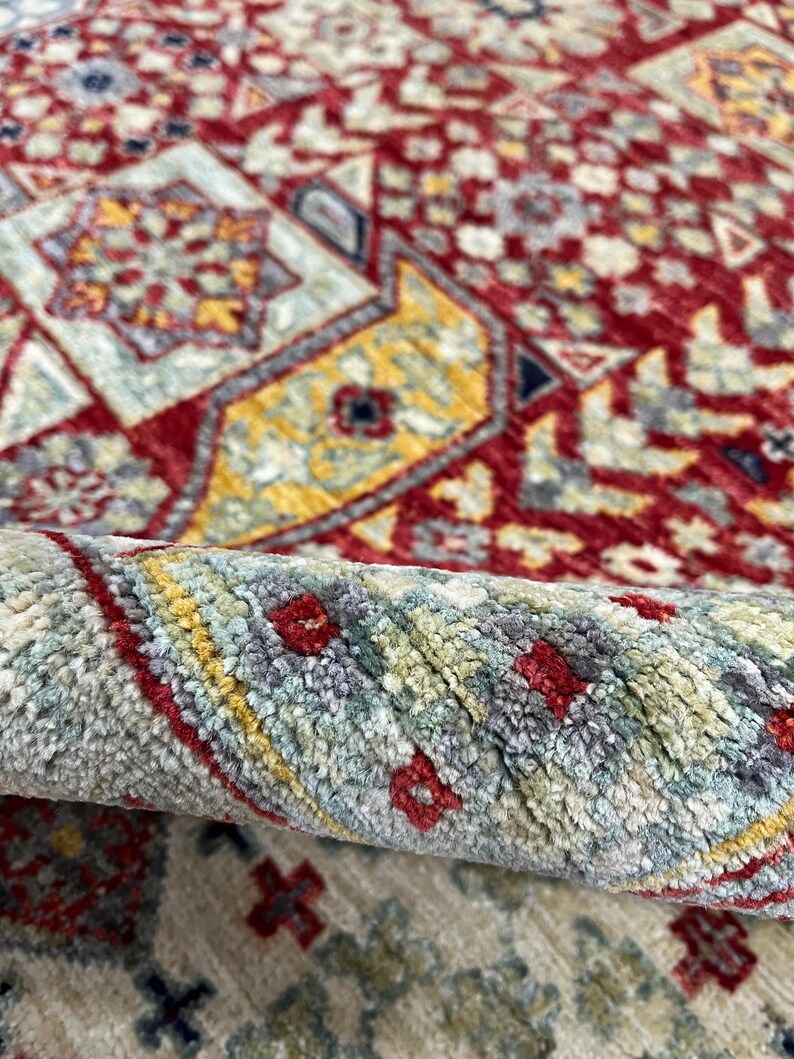 Puede incluir: Primer plano de una alfombra enrollada con un patr&oacute;n intrincado. La alfombra presenta un fondo rojo intenso con dise&ntilde;os geom&eacute;tricos en amarillo, azul y crema. El borde enrollado revela la textura y las variaciones de color.