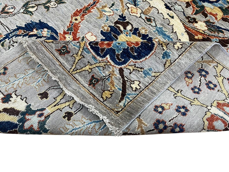 Puede incluir: Primer plano de una alfombra gris con un dise&ntilde;o floral. La alfombra presenta un dise&ntilde;o central con elementos florales azules, rojos y amarillos. Los bordes est&aacute;n doblados, revelando el borde con flecos.