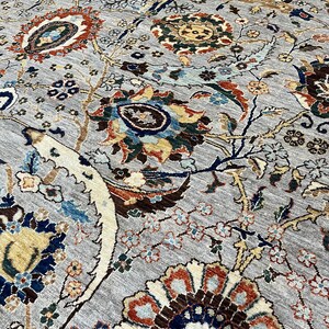 Puede incluir: Primer plano de una alfombra gris con un intrincado dise&ntilde;o floral. El dise&ntilde;o presenta grandes flores estilizadas en tonos marr&oacute;n, azul, naranja y amarillo, sobre un fondo gris. La alfombra tiene una apariencia texturizada, lo que sugiere una construcci&oacute;n tejida.
