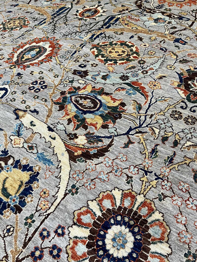 Puede incluir: Primer plano de una alfombra gris con un intrincado dise&ntilde;o floral. El dise&ntilde;o presenta grandes flores estilizadas en tonos marr&oacute;n, azul, naranja y amarillo, sobre un fondo gris. La alfombra tiene una apariencia texturizada, lo que sugiere una construcci&oacute;n tejida.