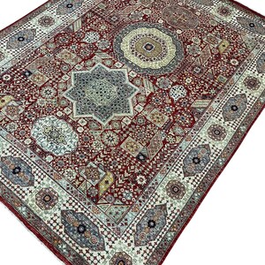 Puede incluir: Una alfombra rectangular detallada con un rico fondo rojo y patrones geom&eacute;tricos intrincados. La alfombra presenta un dise&ntilde;o central en forma de estrella, rodeado de motivos florales y geom&eacute;tricos en tonos de azul, beige y verde. El borde es de un tono m&aacute;s claro con patrones repetidos.