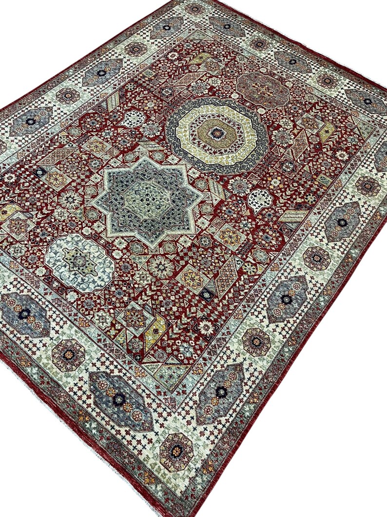 Puede incluir: Una alfombra rectangular detallada con un rico fondo rojo y patrones geom&eacute;tricos intrincados. La alfombra presenta un dise&ntilde;o central en forma de estrella, rodeado de motivos florales y geom&eacute;tricos en tonos de azul, beige y verde. El borde es de un tono m&aacute;s claro con patrones repetidos.
