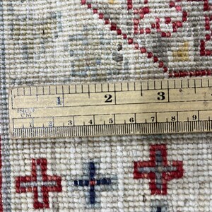 Puede incluir: Primer plano de una alfombra beige y roja estampada con una regla de madera que mide en pulgadas y cent&iacute;metros. La alfombra presenta un dise&ntilde;o de cruces repetidas en rojo y azul.