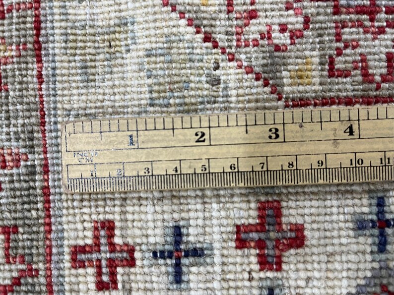 Puede incluir: Primer plano de una alfombra beige y roja estampada con una regla de madera que mide en pulgadas y cent&iacute;metros. La alfombra presenta un dise&ntilde;o de cruces repetidas en rojo y azul.