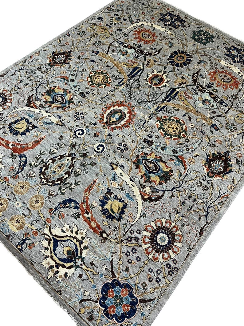 Puede incluir: Una alfombra gris con un intrincado dise&ntilde;o floral. La alfombra presenta un dise&ntilde;o central de flores y hojas estilizadas en tonos de azul, rojo, marr&oacute;n y crema. El borde de la alfombra es de un gris m&aacute;s oscuro.