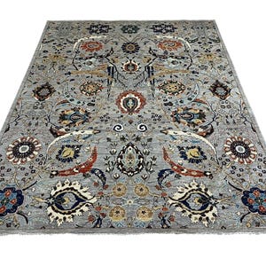 Puede incluir: Una alfombra rectangular grande con un intrincado dise&ntilde;o floral. La alfombra presenta un fondo gris claro con un dise&ntilde;o complejo de flores, hojas y otros elementos decorativos en tonos de azul, rojo y dorado. La alfombra tiene flecos en los bordes.
