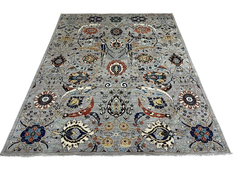 Puede incluir: Una alfombra rectangular grande con un intrincado dise&ntilde;o floral. La alfombra presenta un fondo gris claro con un dise&ntilde;o complejo de flores, hojas y otros elementos decorativos en tonos de azul, rojo y dorado. La alfombra tiene flecos en los bordes.