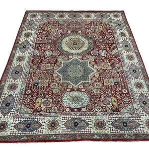 Puede incluir: Una alfombra rectangular grande con un dise&ntilde;o detallado y ornamentado. La alfombra presenta un fondo rojo oscuro con un patr&oacute;n central en forma de estrella en tonos azules y beige. El borde es de un color m&aacute;s claro con motivos geom&eacute;tricos y florales.