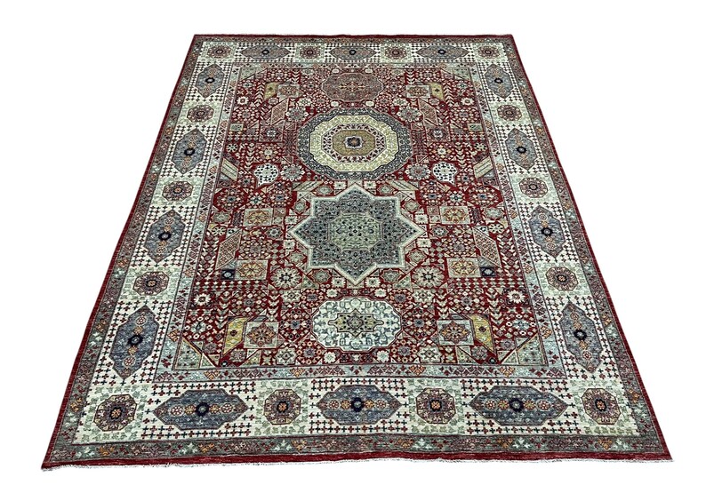 Puede incluir: Una alfombra rectangular grande con un dise&ntilde;o detallado y ornamentado. La alfombra presenta un fondo rojo oscuro con un patr&oacute;n central en forma de estrella en tonos azules y beige. El borde es de un color m&aacute;s claro con motivos geom&eacute;tricos y florales.