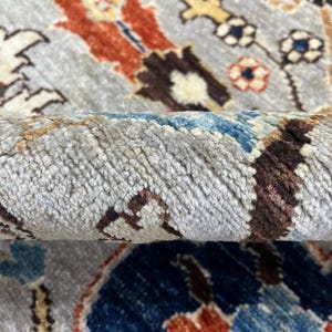 Puede incluir: Primer plano de una alfombra enrollada con un patr&oacute;n intrincado. La alfombra presenta un fondo gris claro con un dise&ntilde;o complejo en tonos de azul, naranja, marr&oacute;n y crema. La textura de la alfombra es visible.