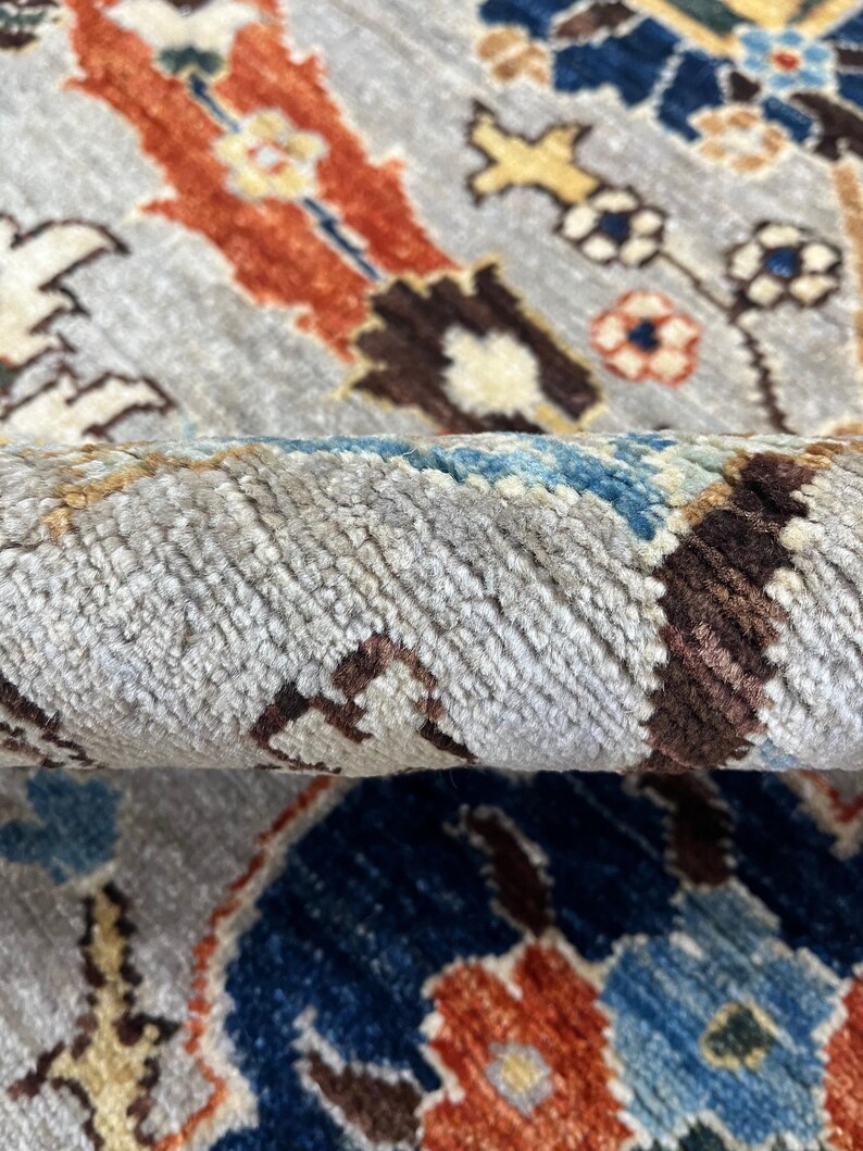 Puede incluir: Primer plano de una alfombra enrollada con un patr&oacute;n intrincado. La alfombra presenta un fondo gris claro con un dise&ntilde;o complejo en tonos de azul, naranja, marr&oacute;n y crema. La textura de la alfombra es visible.