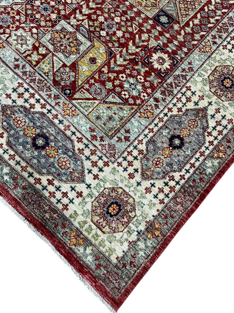 Puede incluir: Primer plano de una secci&oacute;n de una alfombra estampada. La alfombra presenta un dise&ntilde;o geom&eacute;trico complejo con una paleta de colores rojo, beige, gris y amarillo. El borde tiene un patr&oacute;n repetitivo de cruces y oct&aacute;gonos.