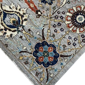 Puede incluir: Primer plano de una alfombra gris con un intrincado dise&ntilde;o floral. El dise&ntilde;o presenta flores y hojas azules, rojas y crema sobre un fondo gris. La alfombra tiene un borde detallado y un borde con flecos.