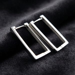 Può includere: Un paio di orecchini a cerchio rettangolari argentati. Gli orecchini sono in metallo lucido e hanno un design semplice e moderno. Sono esposti su una superficie grigio scuro strutturata.