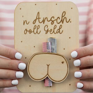 Könnte beinhalten: Ein Geschenkkartenhalter aus Holz mit einem Ausschnitt eines Cartoon-Hinterns und dem deutschen Satz "'n Arsch. voll Geld" (ein Arsch voller Geld). Der Halter hat einen Schlitz für Bargeld und wird von einer Person mit weißem Nagellack gehalten.