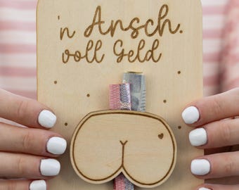 May include: A wooden gift card holder with a cutout of a cartoon butt and the German phrase "'n Arsch. voll Geld" (a** full of money). The holder has a slot for cash and is held by a person with white nail polish.