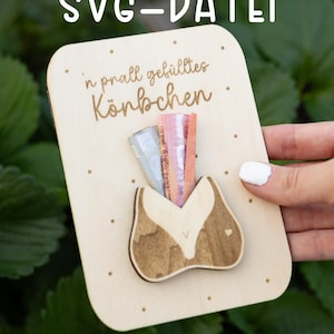 May include: Wooden card with a fox-shaped pocket holding colorful ribbons. The card has the text "SVG-Datei" and "'n prall gefülltes Körbchen" in a handwritten font. Small dots are scattered around the card.