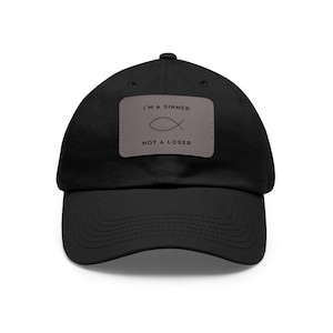 May include: Black baseball cap with a curved brim. A rectangular patch on the front displays the text "I'M A SINNER" above a fish symbol and "NOT A LOSER" in a simple font. The cap is made of a soft fabric.