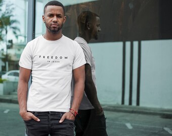 Puede incluir: Camiseta blanca con el texto negro "FREEDOM IN JESUS". La persona lleva vaqueros negros. El texto está centrado en el pecho. El fondo es una pared de cristal.