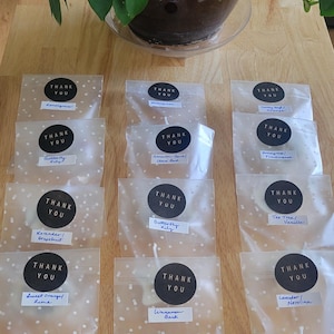 May include: An assortment of small, decorative wax melts in clear cellophane bags, each with a black circular sticker that says "THANK YOU." The melts are various shapes and colors, with handwritten labels indicating scents like Lavender, Sweet Orange, and Cinnamon Bark.