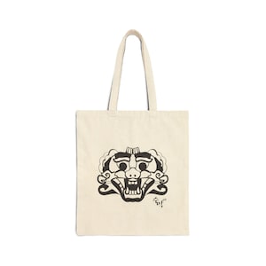 May include: Cream-colored tote bag featuring a black graphic of a traditional Korean mask. The mask has detailed features, including open mouth and stylized eyes. The bag has long handles for carrying.