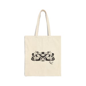 May include: Cream-colored tote bag with a black graphic of a stylized mask. The mask design features intricate details and a smiling mouth. The bag has two handles for carrying and the text "숨막" is printed below the mask.