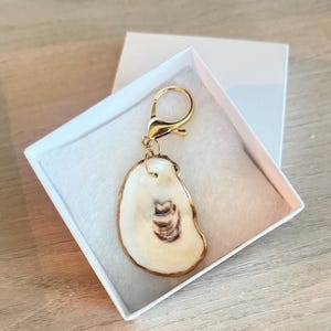 以下が含まれることがあります： 金色の留め金が付いた手作りのカキ殻キーホルダー。 殻は白で、茶色の模様と金色の縁があります。 キーホルダーは、柔らかい白い内装の白いギフトボックスに入っています。