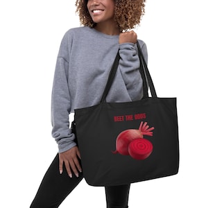 May include: Black tote bag featuring a graphic of two beets, one whole and one sliced, with the text "BEET THE ODDS" in red. The bag has long black handles and is held by a person.