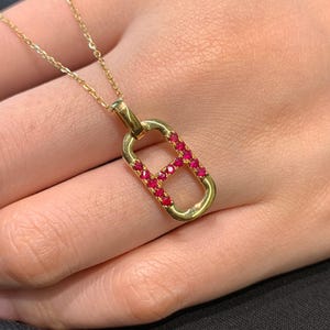 Puede incluir: Un collar de oro con un colgante en forma de letra "B" con piedras preciosas rojas.