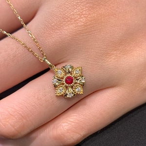 Könnte beinhalten: Eine goldene Halskette mit einem Anhänger in Form einer Blume, der mit einem roten Edelstein verziert ist, der von kleineren weißen Edelsteinen umgeben ist.