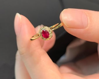 Pode incluir: Um anel de ouro com uma gema oval de rubi vermelho cercada por diamantes menores. O anel é engastado em uma banda torcida delicada.