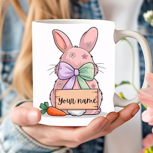 May include: White ceramic mug with a pink bunny design, a purple and green bow, and the text "your name" on a wooden sign. The bunny has small flower details and a carrot. The mug has a white handle.