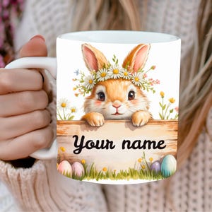 Peut inclure: Mug en céramique blanche avec un lapin de dessin animé portant une couronne de fleurs et tenant une pancarte en bois avec l'inscription "Your name". Le mug a un thème printanier avec des œufs de Pâques et des fleurs.
