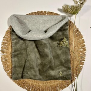 Könnte beinhalten: Ein grüner Cord-Halswärmer mit grauem Fleece-Futter. Der Halswärmer liegt auf einer geflochtenen Strohmatte.