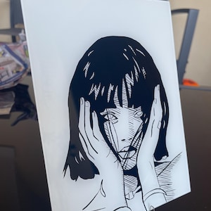 Può includere: Un'opera d'arte rettangolare in vetro bianco con un'illustrazione in bianco e nero di una donna che si tiene il viso. L'opera d'arte ha uno stile pop art con linee e ombreggiature audaci. Il pezzo è esposto su un supporto trasparente.
