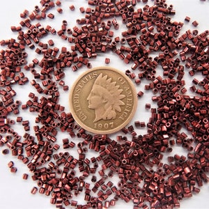 May include: A close-up of a pile of small, red, metallic beads scattered on a white surface. A copper-colored US penny coin with the inscription "UNITED STATES OF AMERICA" and the year "1907" is in the center of the image.