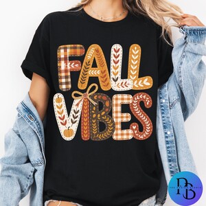 May include: Black t-shirt with the words "FALL VIBES" in a fall-themed design. The letters are decorated with plaid, leaves, and a pumpkin. The shirt is a casual, short-sleeved top.