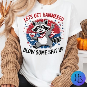May include: White t-shirt with a graphic of a raccoon holding a drink, wearing an American flag bandana, and giving a peace sign. The text reads "LETS GET HAMMERED" and "BLOW SOME SHIT UP" in red and black.