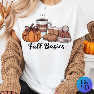 May include: White t-shirt with a fall-themed graphic. The design includes a pumpkin, coffee mug, boots, and a beanie. The words "Fall Basics" are printed below the illustrations. The shirt is made of a soft material.