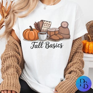 May include: White t-shirt with a graphic of fall essentials: a pumpkin, coffee cup, boots, hat, and scarf. The words "Fall Basics" are printed in black script below the illustrations. The shirt is worn by a person.
