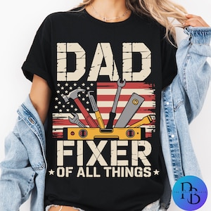 Könnte beinhalten: Schwarzes T-Shirt mit den weißen Worten "DAD FIXER OF ALL THINGS". Das Design beinhaltet eine Grafik von Werkzeugen und eine amerikanische Flagge. Das Shirt hat einen Rundhalsausschnitt.