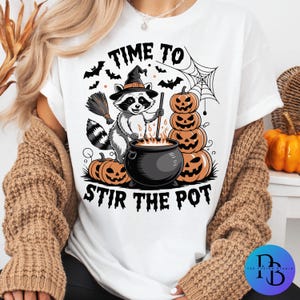May include: White t-shirt with a Halloween graphic. The design shows a raccoon in a witch hat stirring a cauldron, with pumpkins, bats, and the text "TIME TO STIR THE POT". A broom and spiderweb are also included.