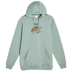 May include: A light green hooded sweatshirt with a pocket on the front. The sweatshirt has an embroidered image of a turtle with the word "Doc" above it.