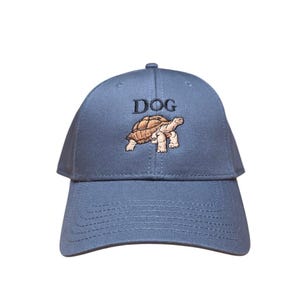 Pode incluir: Boné de beisebol azul com a palavra "DOG" impressa na frente. Abaixo, uma imagem bordada de uma tartaruga. O boné tem uma aba curva e um botão na parte superior.