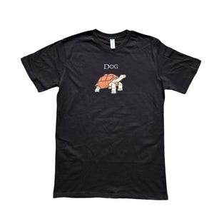 May include: Black t-shirt featuring a cartoon turtle with an orange shell and white legs. The word "Dog" is printed above the turtle. The shirt is a classic crew neck style.
